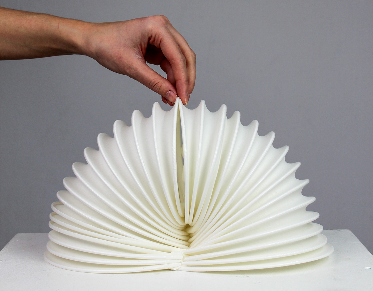 A hand lifting a white 3D-printed textile fan structure that holds its own pleated shape.