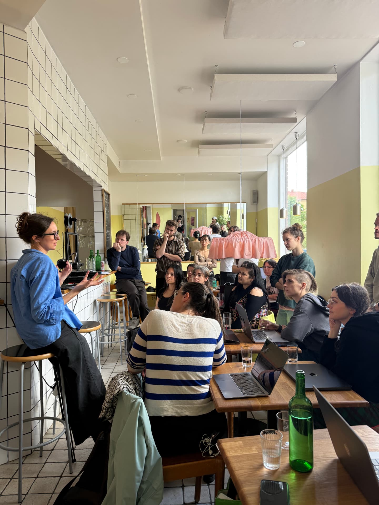 A workshop leader presents to a seated group in a bright tiled room with laptops and notebooks.
